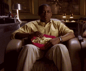 Video gif. A man in a yellow shirt sits on a brown leather armchair, snacking from a bowl of popcorn. We cut to a closeup, where he gives us an interested bob of his head and eats some more popcorn.